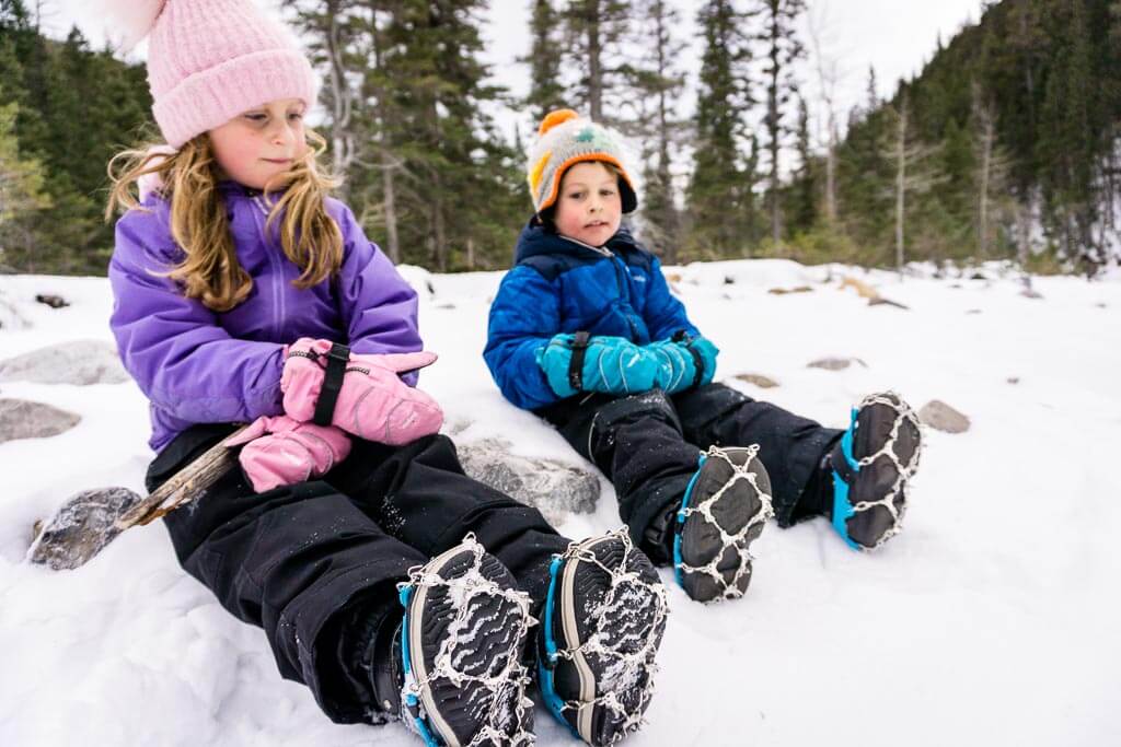 kids sitting on snow with traction devices over their boots.