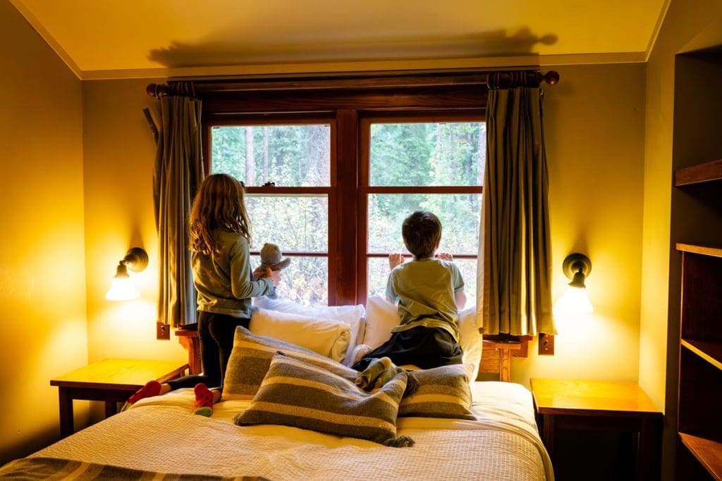 Two kids sitting on a bed look out the window at the Johnston Canyon Lodge and Bungalows.
